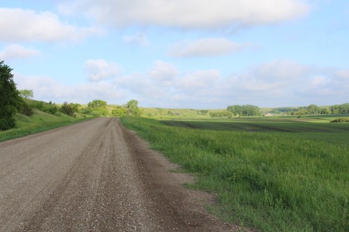 A gravel road stretches through lush green fields under a partly cloudy sky.