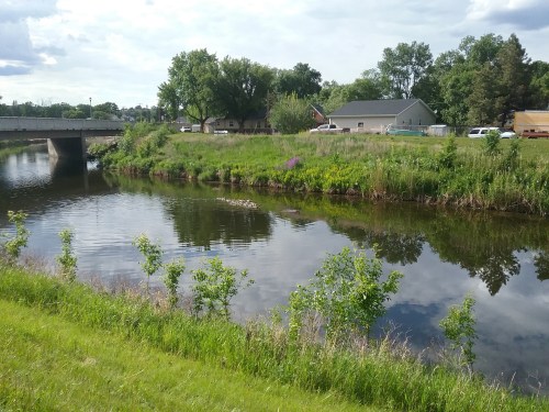 A calm river reflects trees and a building, with green grass and shrubs lining the bank under a partly cloudy sky.