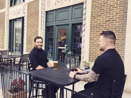 Two men sit at a table outside a café, enjoying drinks and chatting on a sunny day.