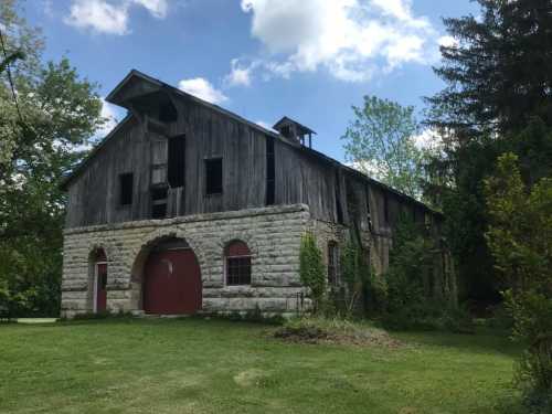 A weathered barn with a stone base and wooden upper structure, surrounded by greenery and under a partly cloudy sky.