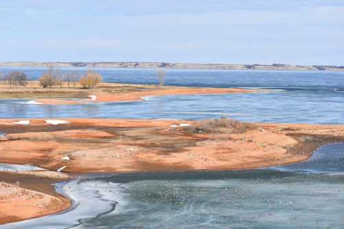 A scenic view of a lake with sandy shores, patches of ice, and distant trees under a clear blue sky.