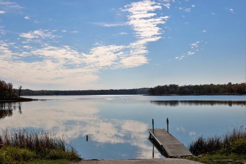 A serene lake scene with a wooden dock, reflecting clouds and trees under a clear blue sky.