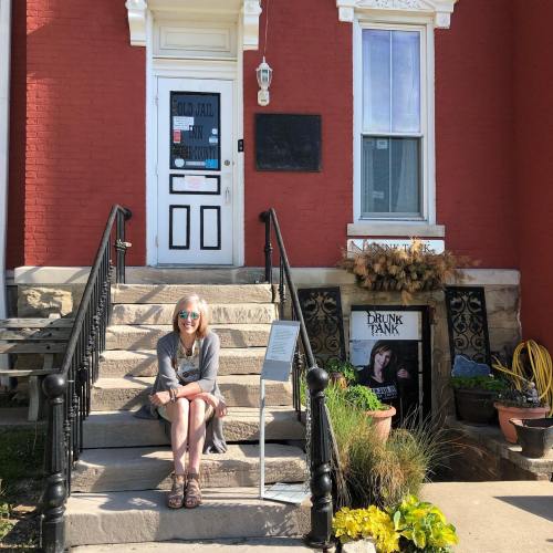 A woman sits on the steps of a red brick building, surrounded by potted plants and signs for nearby businesses.