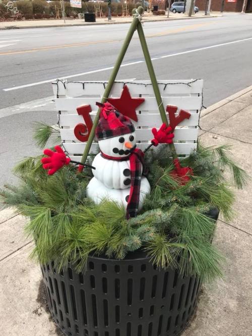A festive snowman in a planter, surrounded by greenery, with a sign that reads "Joy" and wearing a red hat and scarf.