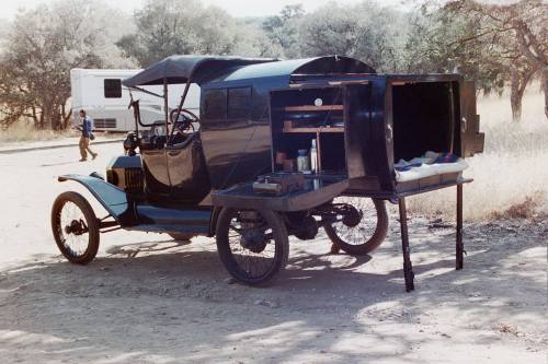 A vintage black car with an open side revealing storage, parked on a dirt road surrounded by trees.