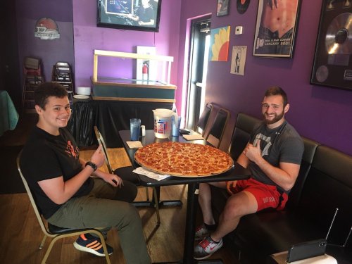 Two young men sit at a table in a colorful restaurant, smiling and giving thumbs up next to a large pizza.