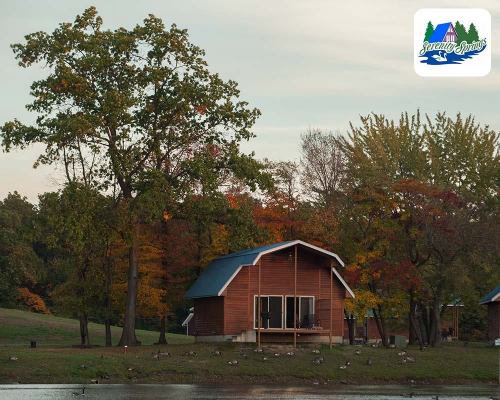 A cozy cabin by a lake surrounded by colorful autumn trees and a clear sky.