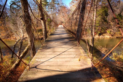 A wooden suspension bridge stretches over a calm river, surrounded by trees in a serene, autumn landscape.