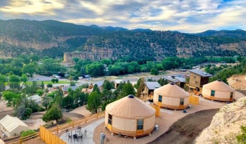 Scenic view of yurts on a hillside, surrounded by trees and mountains under a cloudy sky.