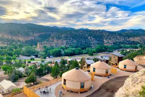 Scenic view of yurts on a hillside, surrounded by trees and mountains under a cloudy sky.