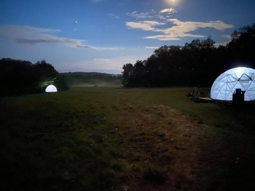 Two illuminated geodesic domes in a dark field under a night sky with clouds and distant hills.