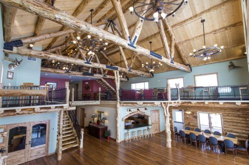 Interior of a spacious lodge with wooden beams, chandeliers, and a staircase, featuring a dining area and seating.