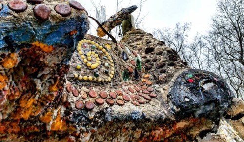 Colorful, textured rock sculpture adorned with various stones and shells, set against a cloudy sky and bare trees.