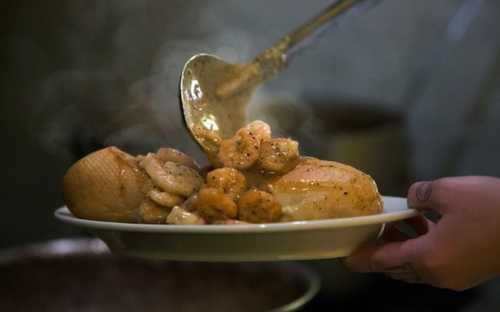 A hand serves a plate of shrimp and chicken, steaming and garnished, from a kitchen setting.