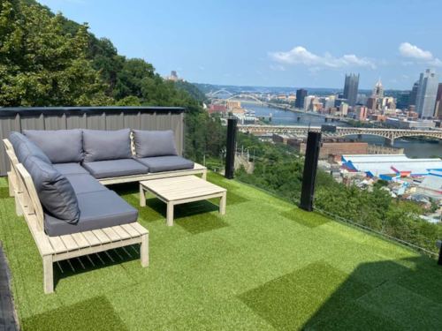 A rooftop patio with a gray sectional sofa and a small table, overlooking a city skyline and river.