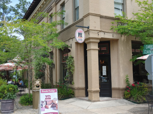 A charming corner building with a sign for "Fresh Fudge," surrounded by greenery and outdoor seating.
