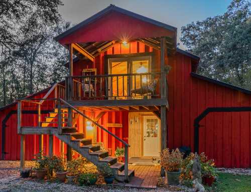 A charming red house with a balcony, surrounded by trees and potted plants, illuminated in the evening light.