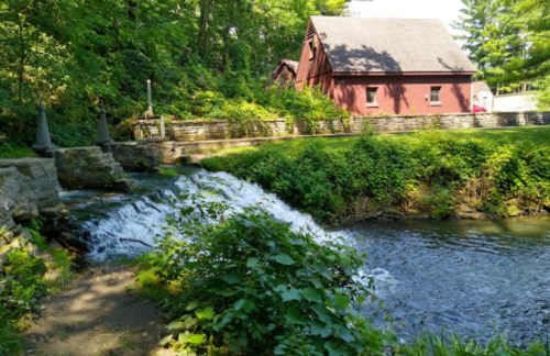 A serene scene featuring a small waterfall flowing into a river, with a red barn and lush greenery in the background.