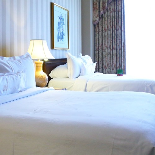 A cozy hotel room featuring two beds, a lamp, and floral curtains with a window letting in natural light.