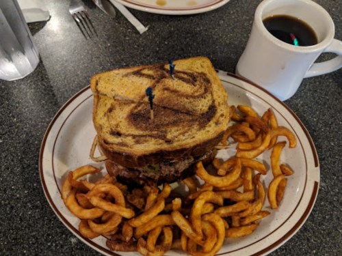 A towering sandwich on a plate, surrounded by curly fries, with a cup of coffee in the background.
