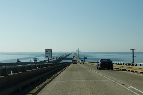 A long bridge stretches over calm water, with vehicles driving and a sign that reads "PASS WITH CARE."