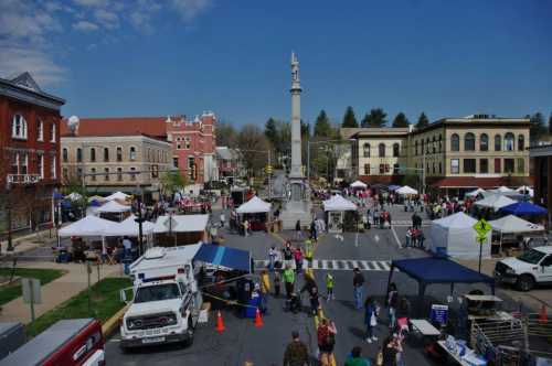 A bustling town square with tents, vendors, and a monument, filled with people enjoying a sunny day.