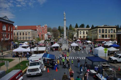 A bustling town square with tents, vendors, and a monument, filled with people enjoying a sunny day.