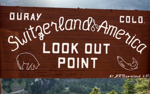 Wooden sign reading "Switzerland of America Look Out Point" with bear and fish illustrations, surrounded by mountains.