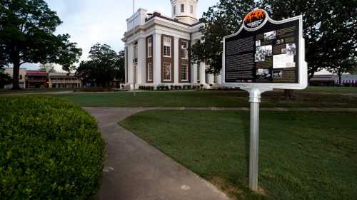 Historic building with a sign detailing local history, surrounded by green grass and trees. Pathway leads to the structure.