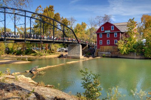 A scenic view of a bridge over a river, with a red building and autumn trees in the background.
