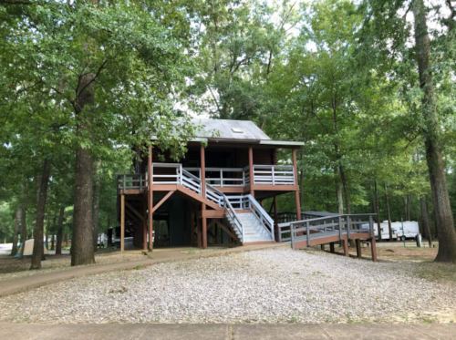 A wooden house on stilts surrounded by trees, with a gravel path leading up to it and a ramp for accessibility.