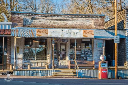 A vintage storefront with a Coca-Cola sign, wooden steps, and a gas pump outside, surrounded by trees.