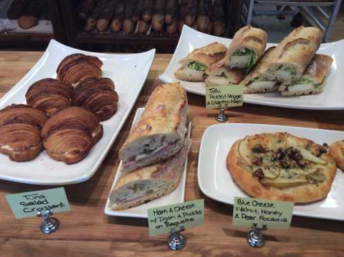 A display of various baked goods: croissants, sandwiches, and a flatbread with toppings, all labeled on a wooden table.