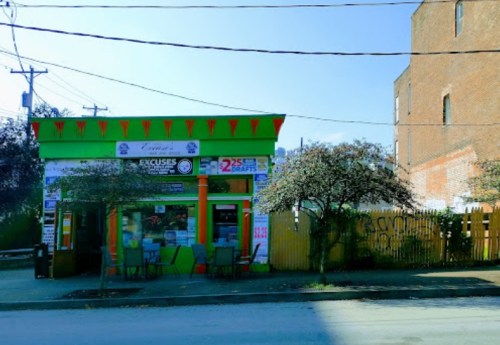 Colorful storefront with green and orange accents, outdoor seating, and trees in front, set against a clear blue sky.