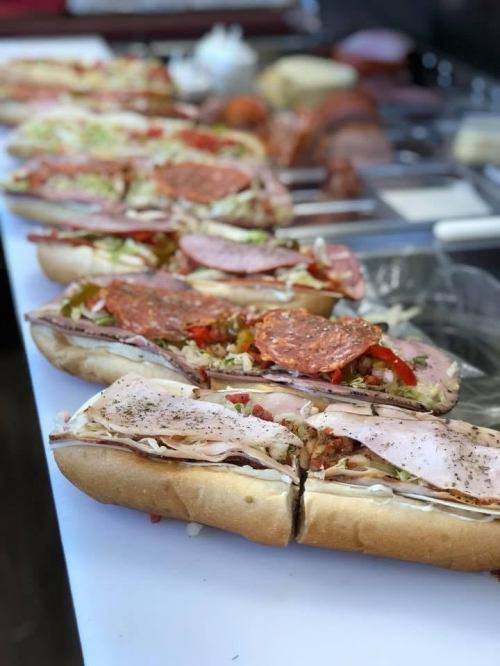 A row of large sandwiches filled with various meats, vegetables, and condiments on a white countertop.