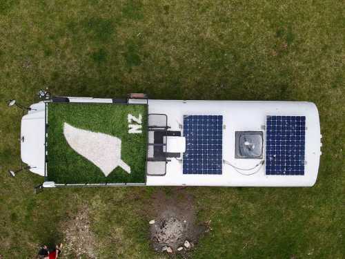 Aerial view of a white RV with solar panels and a green grass patch featuring a leaf and "NZ" on the roof.