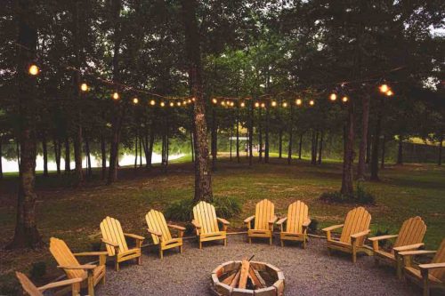 A cozy outdoor fire pit surrounded by wooden chairs, with string lights hanging above and trees in the background.