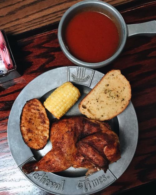 A metal plate with a roasted chicken leg, corn on the cob, garlic bread, and a bowl of tomato soup.