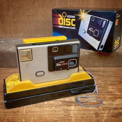 A Kodak Disc 4000 camera on a yellow base, next to its original box, set against a wooden background.