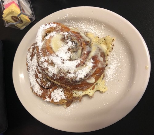 A cinnamon roll topped with icing and powdered sugar, served on a plate with a waffle underneath.