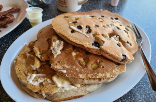 A plate of thick pancakes with blueberries and butter, served with a side of syrup.