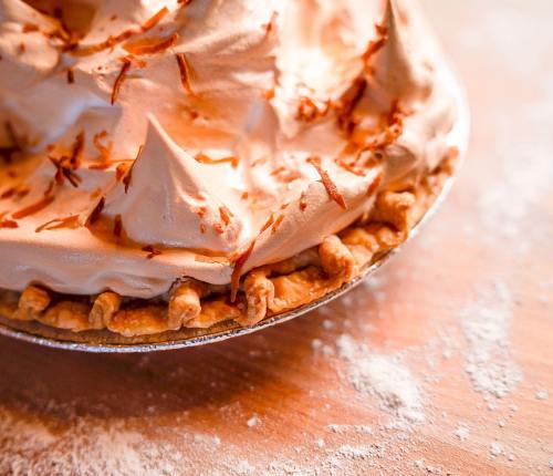 A close-up of a meringue-topped pie with a golden crust, sprinkled with toasted coconut flakes on a wooden surface.