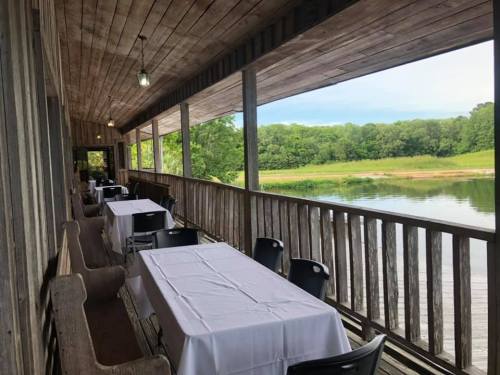 A rustic wooden porch with tables overlooking a calm river and green landscape.