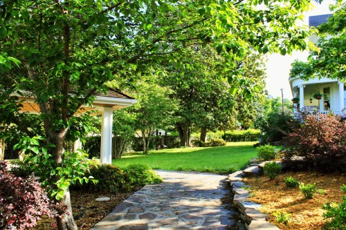 A serene garden path lined with trees and shrubs, leading to a lush green lawn and a charming house in the background.