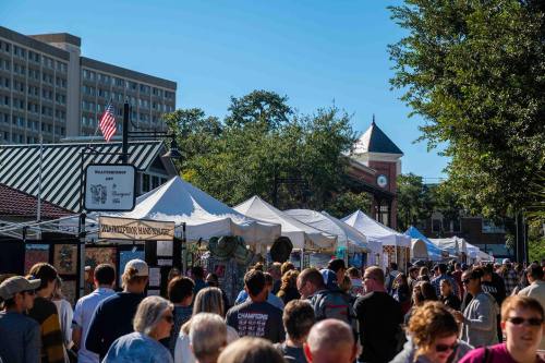 A bustling outdoor market with white tents, people browsing, and a clock tower in the background on a sunny day.