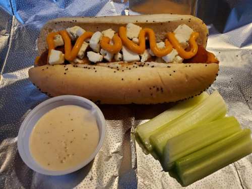 A hot dog topped with cheese and mustard, served with celery sticks and a side of dipping sauce on a silver foil background.