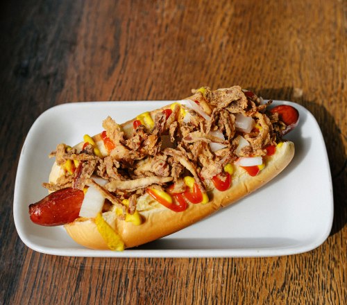 A hot dog topped with fried onions, mustard, and ketchup on a white plate, set on a wooden table.