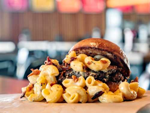 A close-up of a burger topped with macaroni and cheese, bacon, served on a wooden table in a casual dining setting.