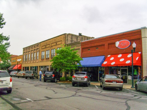 A street view of a small town with shops, parked cars, and trees under a cloudy sky.