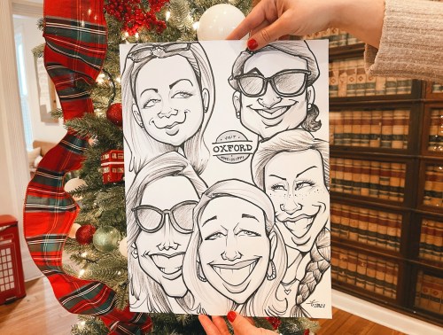 A hand holds a caricature drawing of five smiling women in front of a decorated Christmas tree.